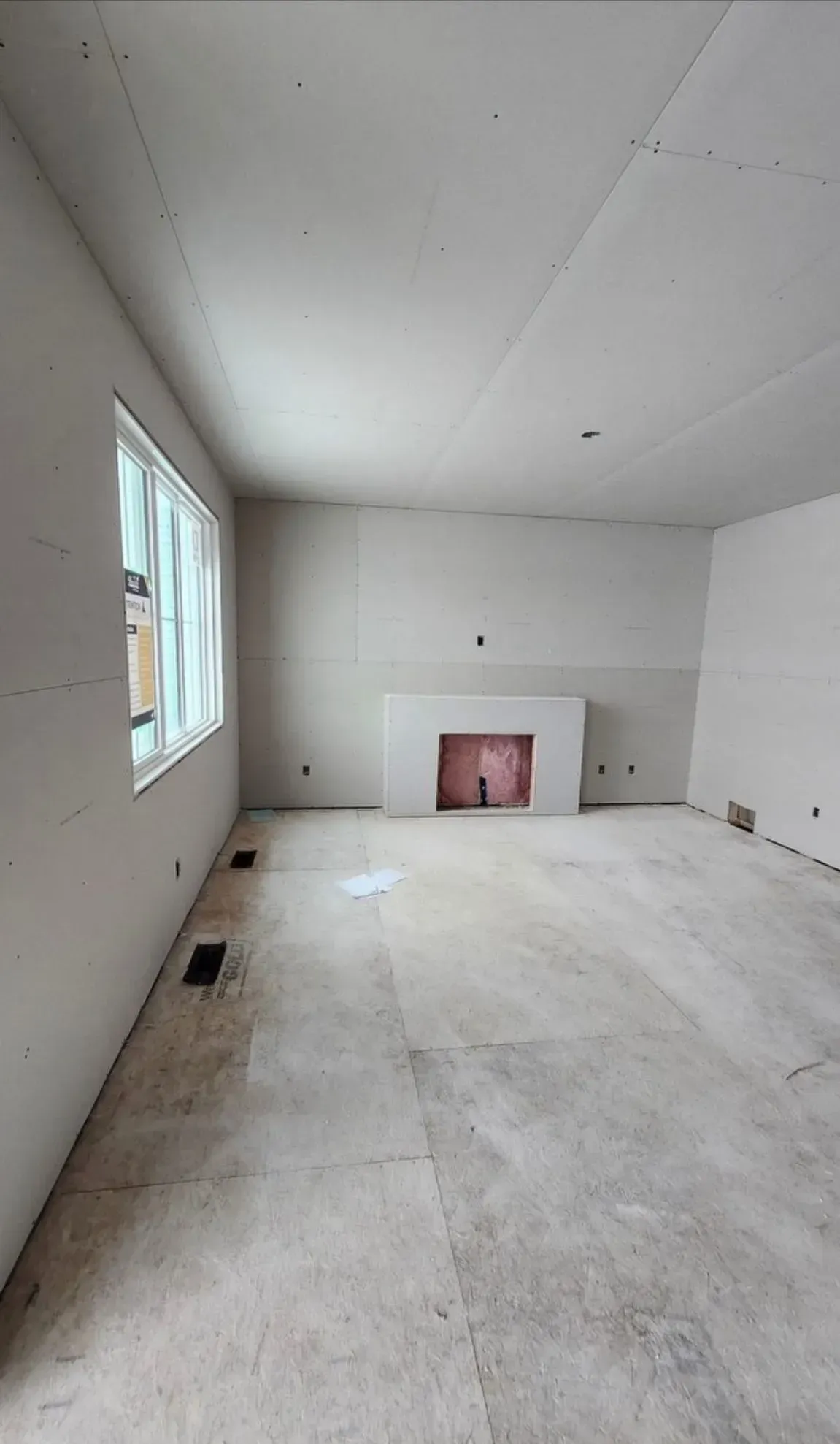 Empty white room with a window, bare floor, and a fireplace opening on the far wall