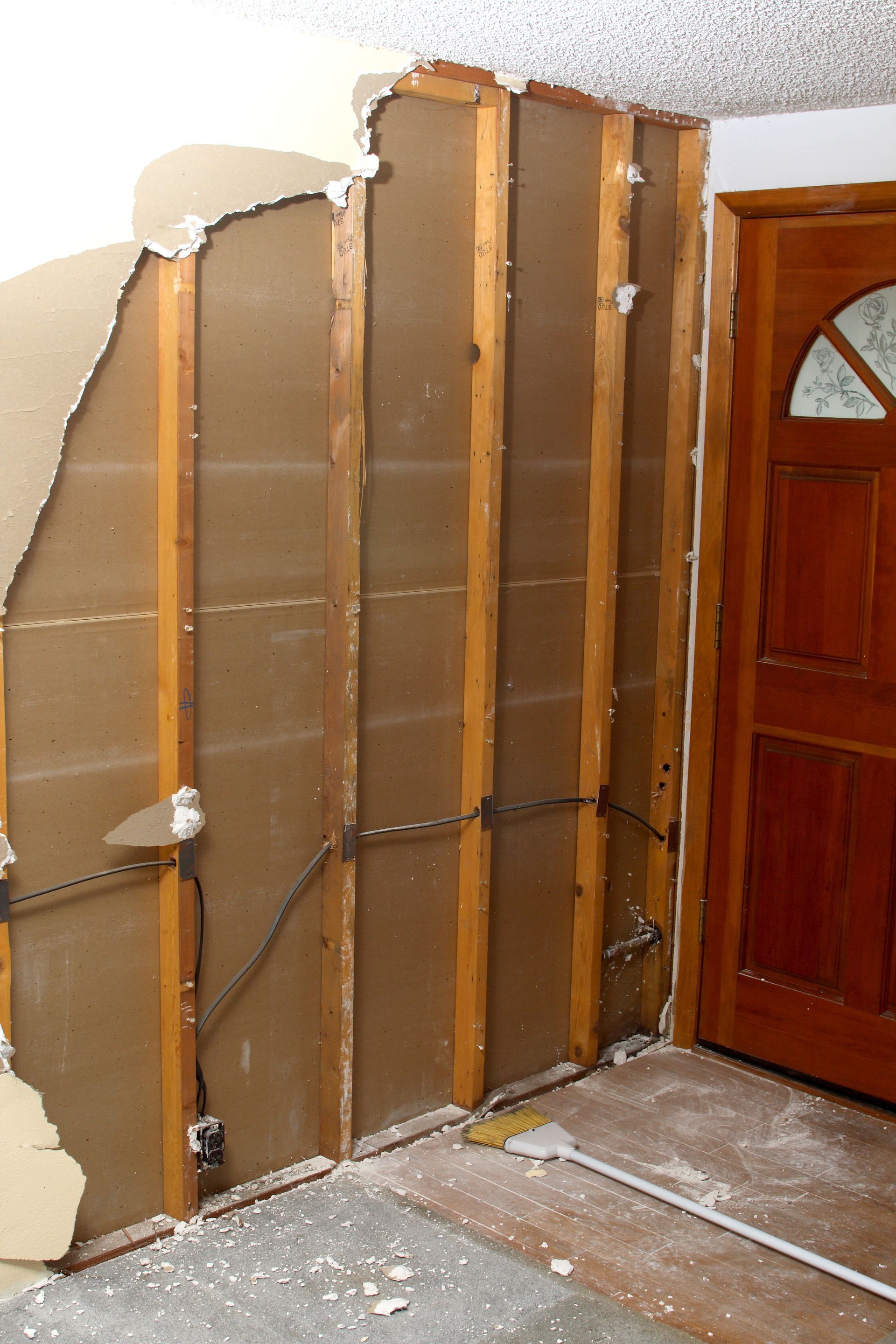 Damaged interior wall with exposed studs and insulation next to a wooden door and dusty floor