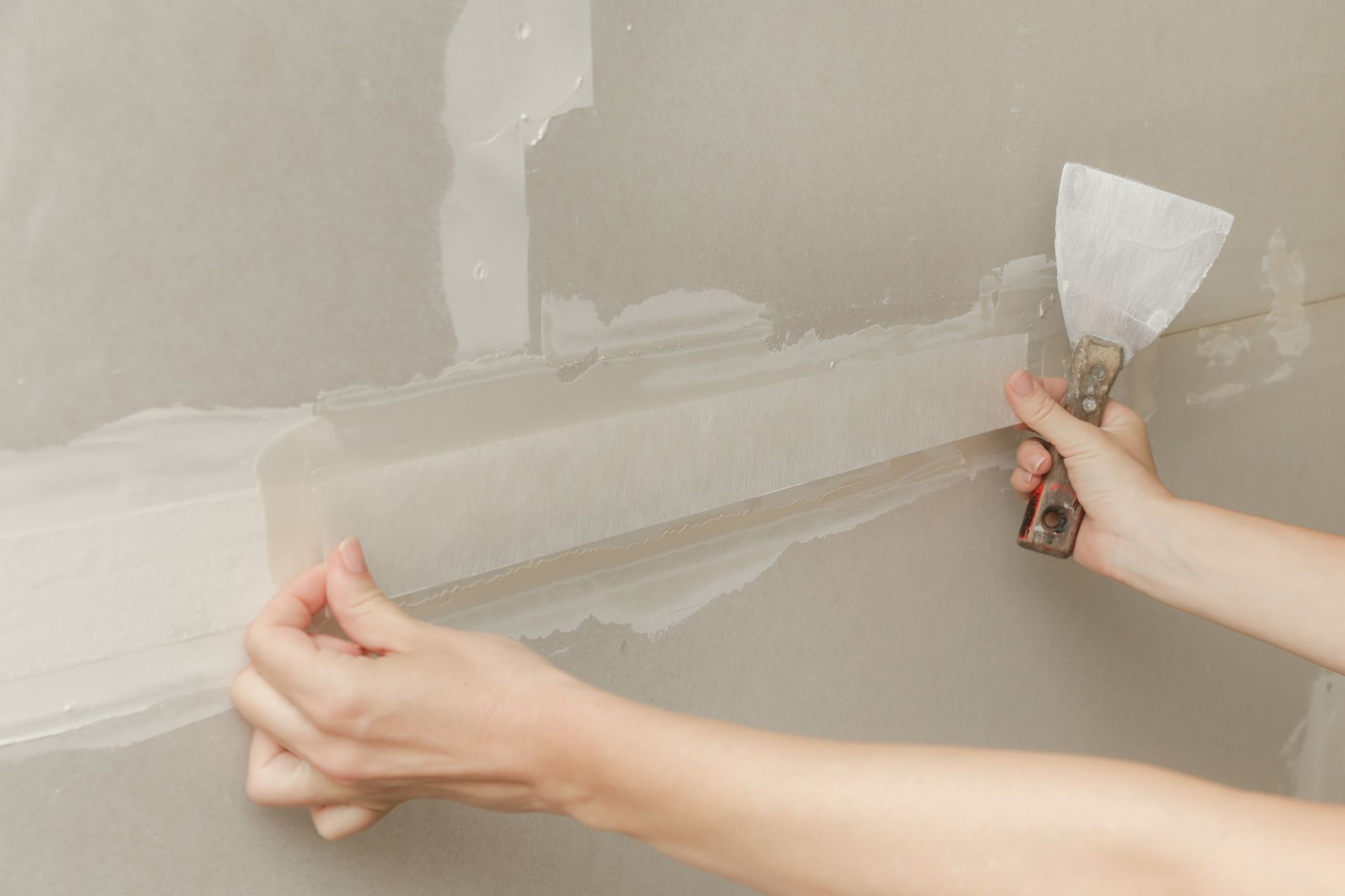 Hands spreading white joint compound on a gray wall with a putty knife