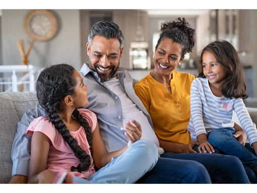 A family of four smiling and interacting while sitting together on a comfortable sofa in a bright, modern living room.