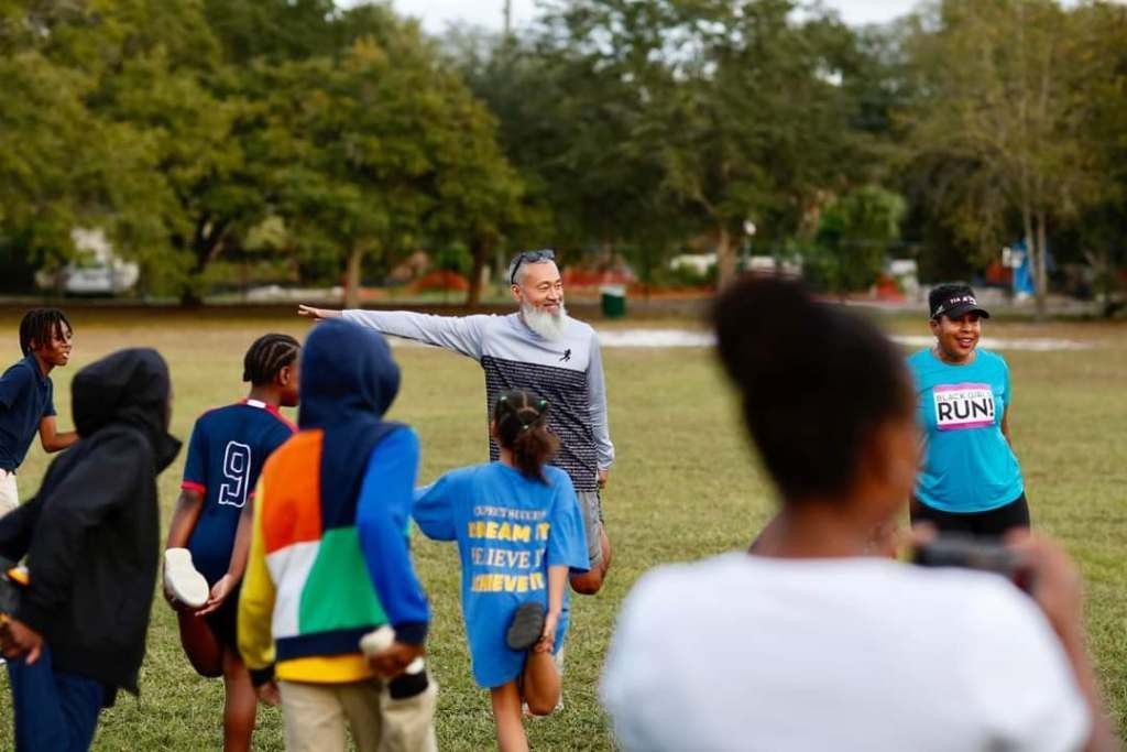 A bearded coach points across a grassy field to a group of people in athletic wear during an outdoor practice session.