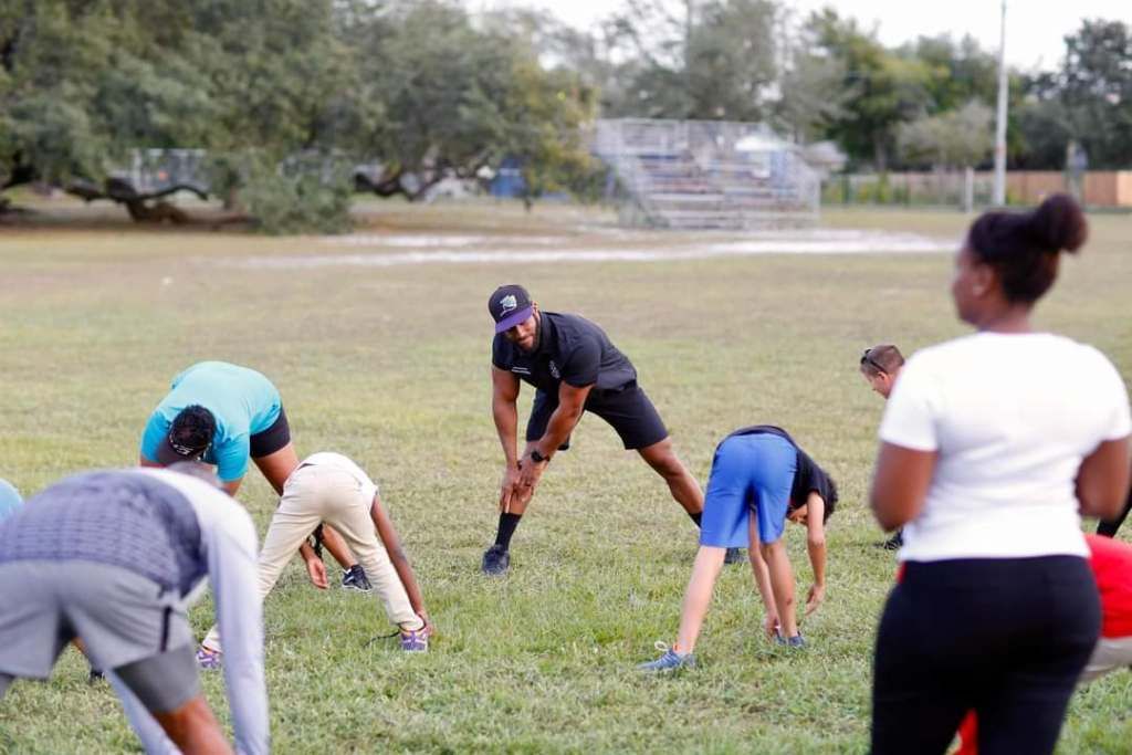 A group of people performs stretches on a grass field under the instruction of a person standing in the center.