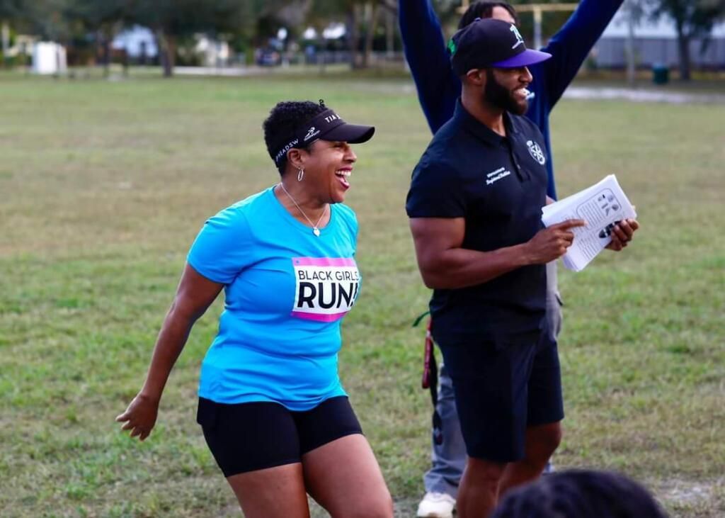 A smiling person in a blue shirt and visor walks beside someone in a black polo and cap holding papers in a grassy field.