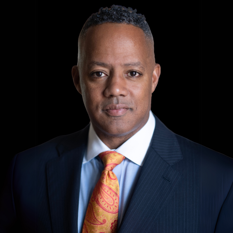 Mark a. board member. A person wearing a dark suit, light shirt, and patterned orange tie against a solid black background.