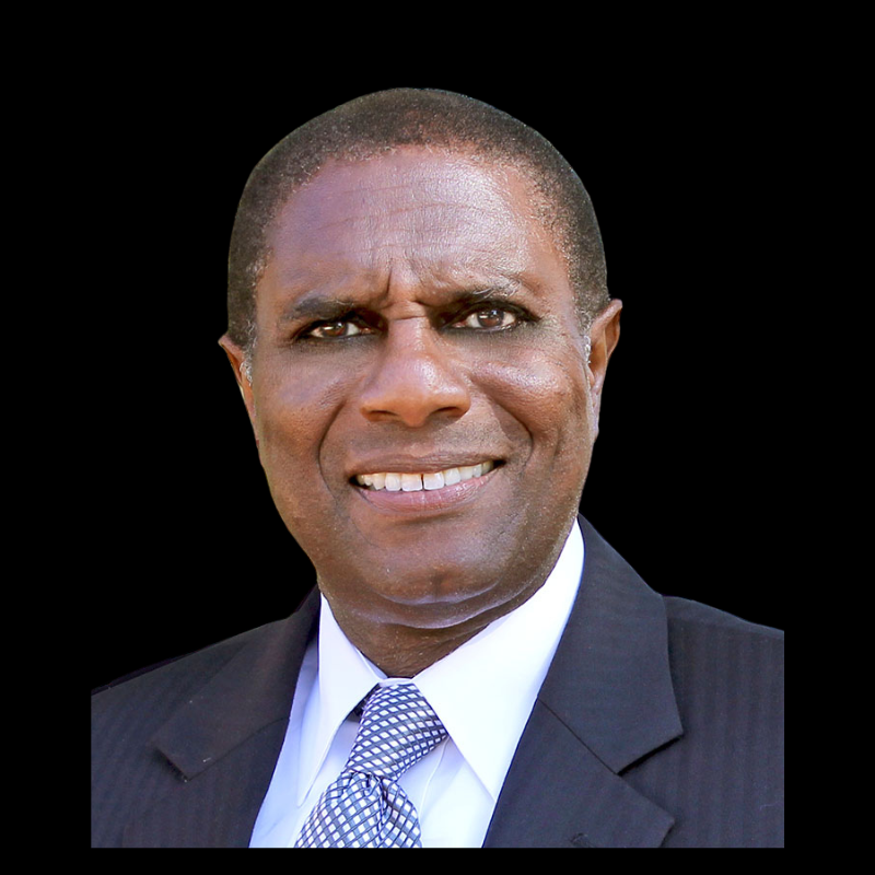 Tony a board member Headshot of a man smiling, wearing a dark suit, light shirt, and patterned tie against a black background.