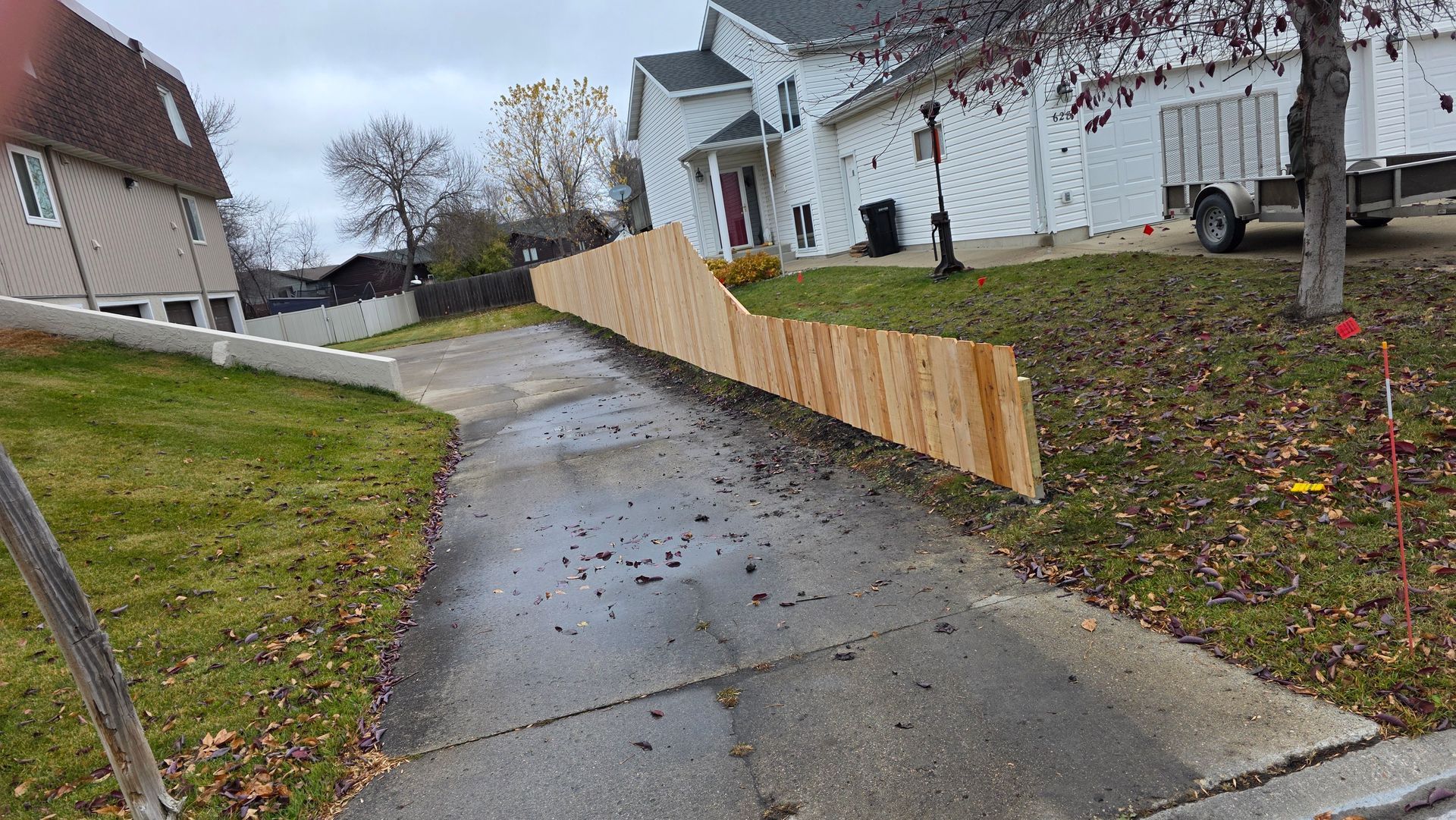 Wooden fence on a slope alongside a driveway, with houses and overcast sky in the background.