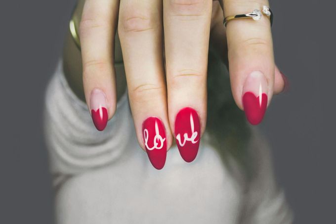 A close-up of a hand with red, heart-shaped French manicure nails featuring white cursive letters spelling out 