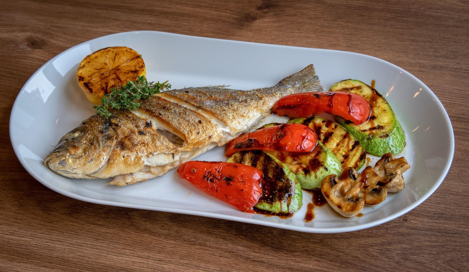 Pesce alla griglia con verdure arrosto su un piatto bianco, su un tavolo di legno