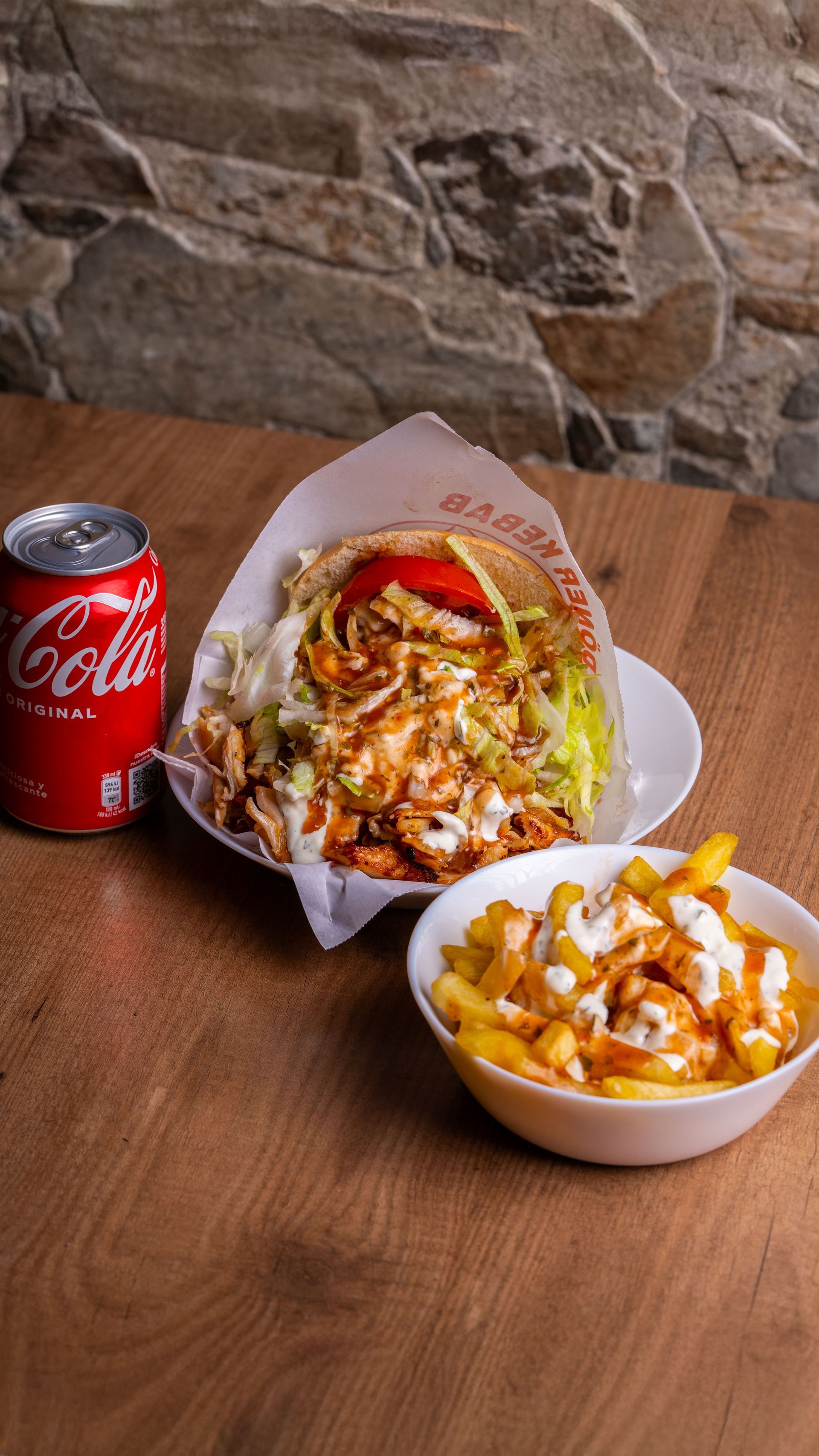 Lattina di Coca-Cola accanto a un wrap e a una ciotola di patatine fritte su un tavolo di legno, con una parete in pietra sullo sfondo