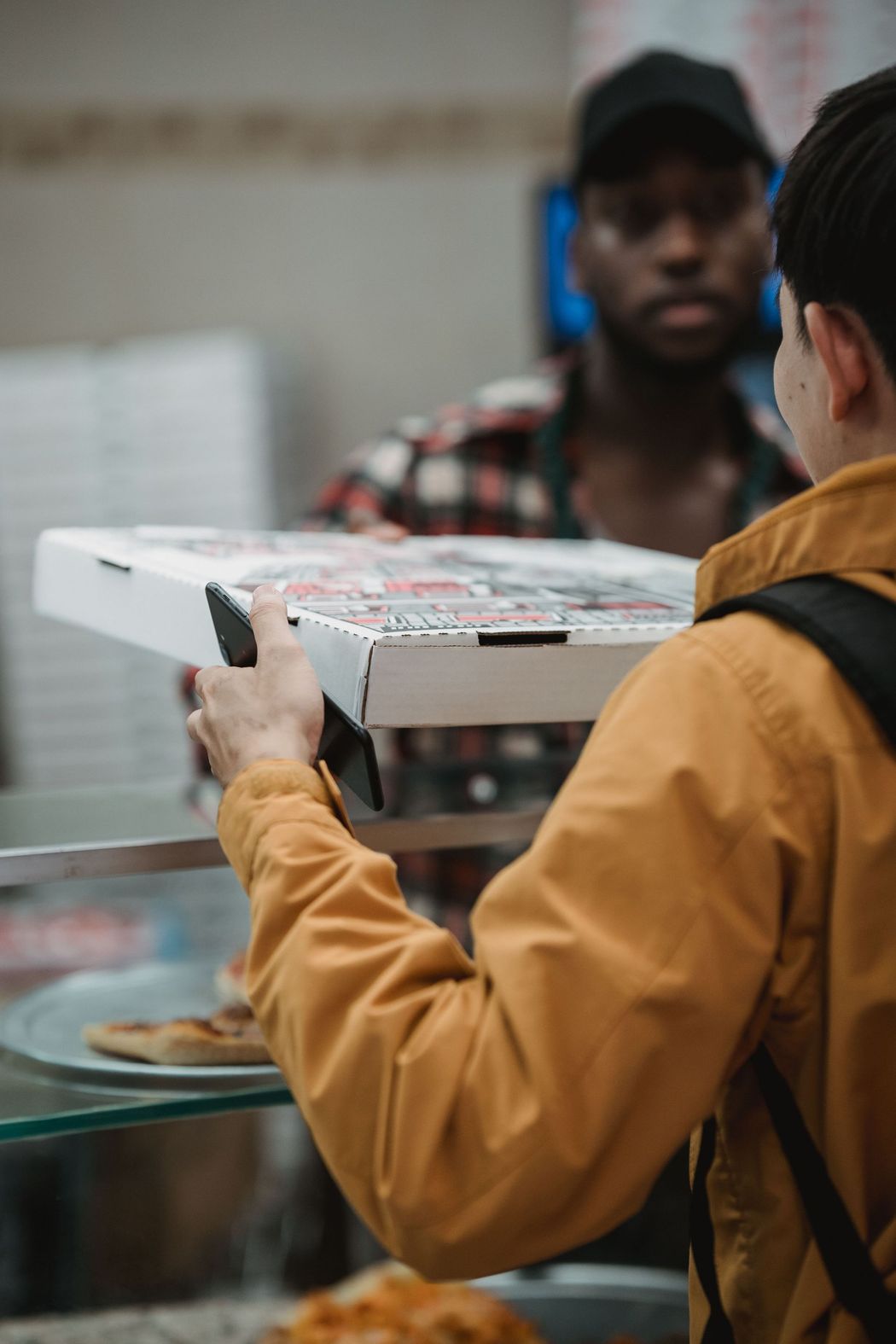 Una persona con una giacca color senape passa una scatola della pizza oltre il bancone a un addetto dietro un vetro