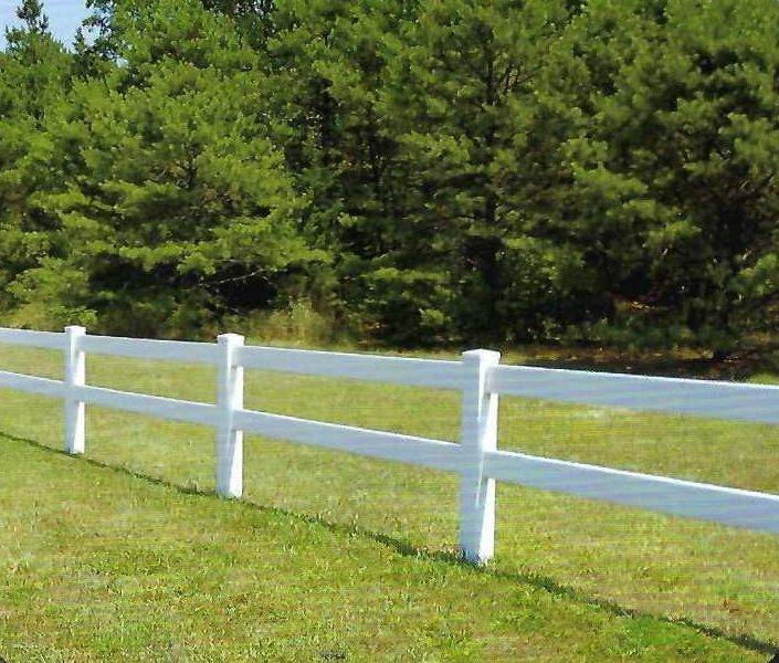 White two-rail fence in a grassy field with a backdrop of green trees.