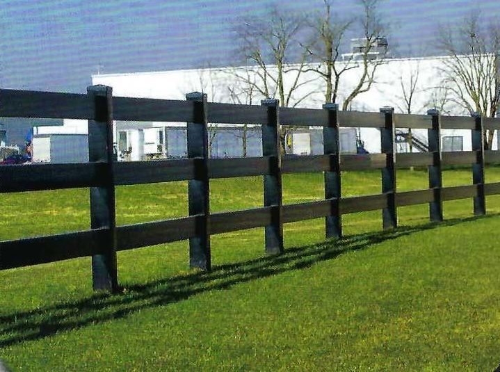 A brown, three-rail fence on a grassy field with a building in the background.