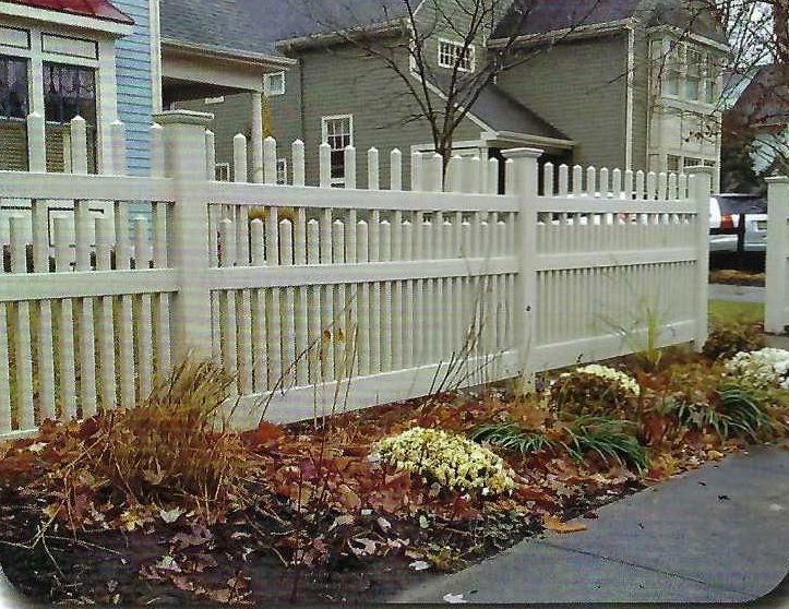 White picket fence bordering a garden with brown mulch and green plants. Houses in background.