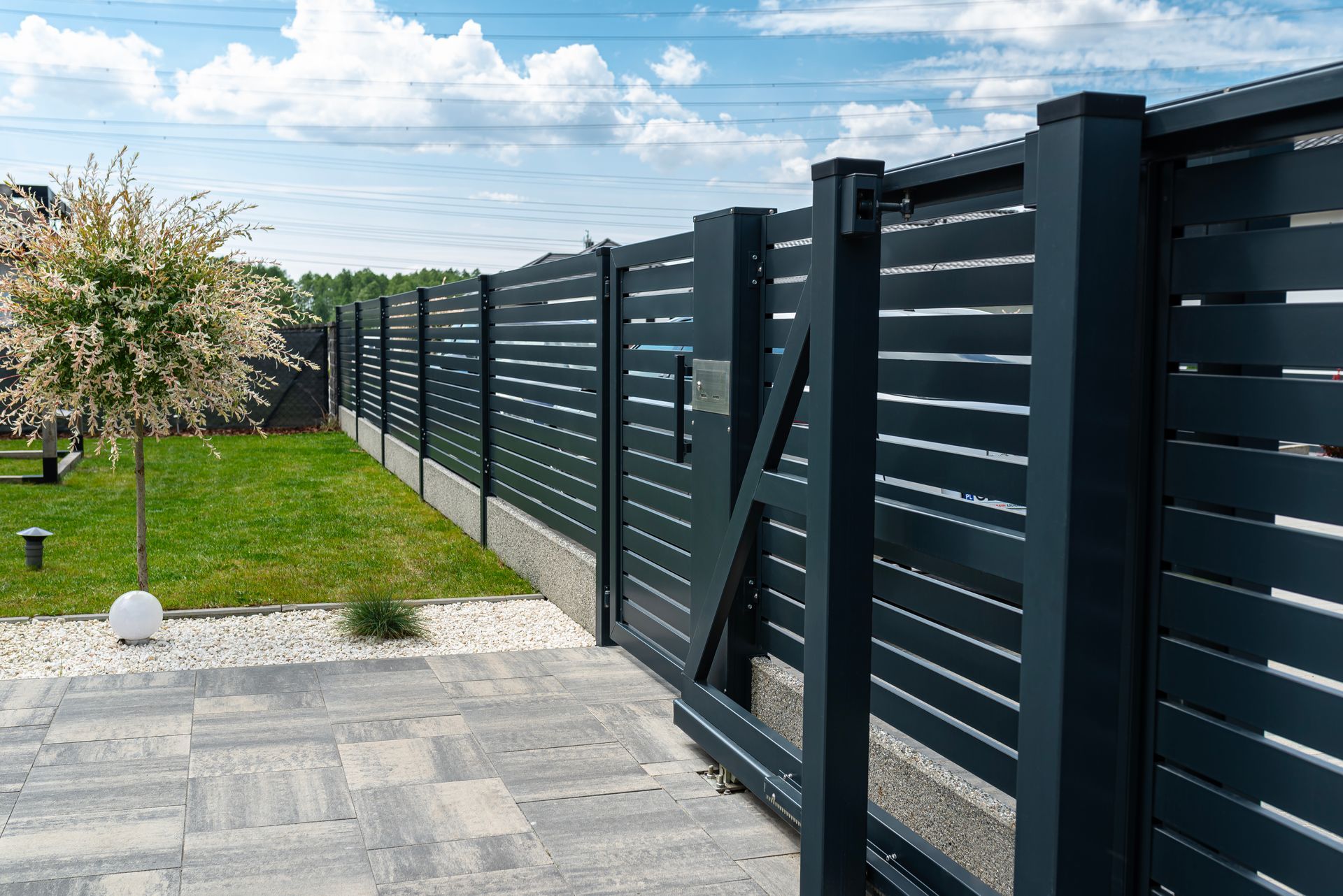 Modern black metal fence along a paved walkway beside a grassy yard and tree in Central PA under a cloudy sky.