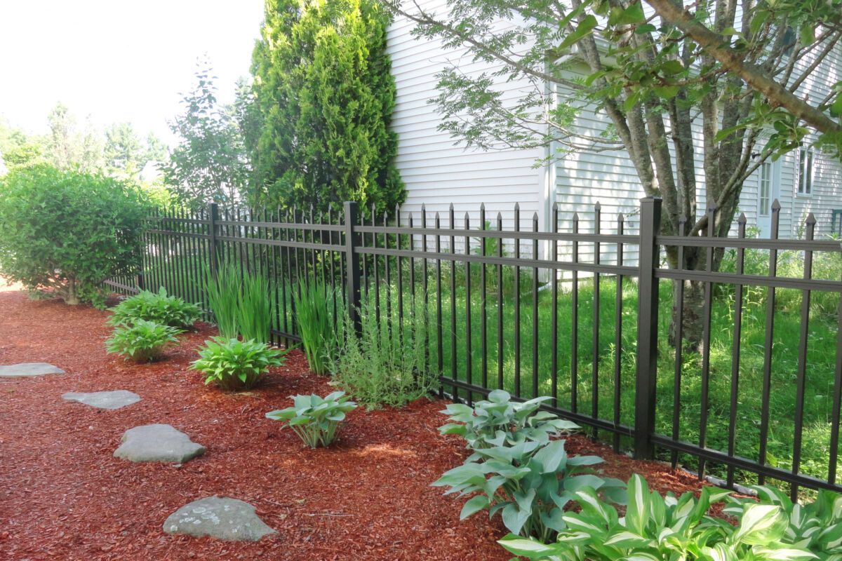 Landscaped garden with red mulch, stepping stones, shrubs, and a black metal fence beside a house