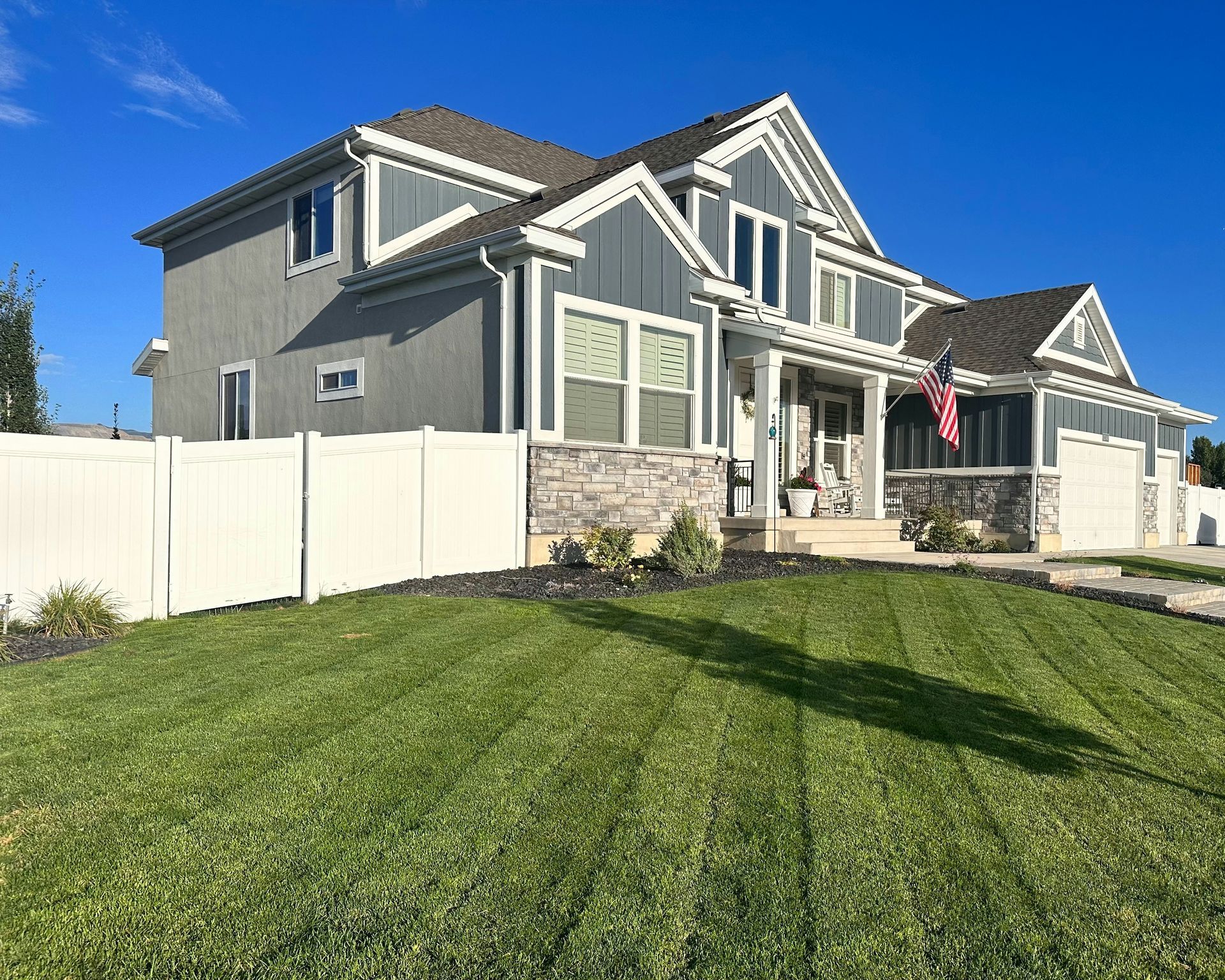 Two-story gray suburban house with white trim, stone porch, white fence, and a green lawn under a blue sky
