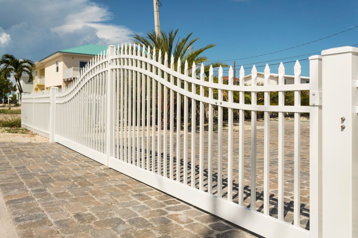 White arched metal driveway gate beside a stone-paved path under a clear blue sky