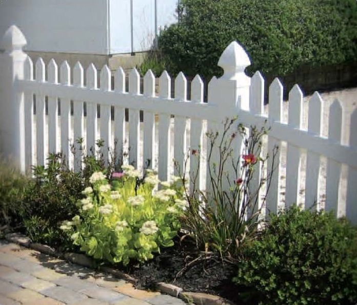 White picket fence with flowers and bushes in a garden bed next to a brick walkway.