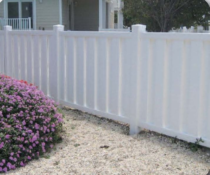 White vinyl fence encloses a yard with a small gravel area and a bush with purple flowers.