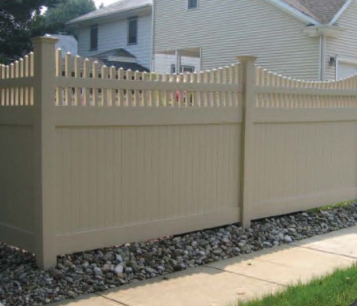 Tan vinyl fence with decorative top, separating a yard from a sidewalk, with stone ground cover.
