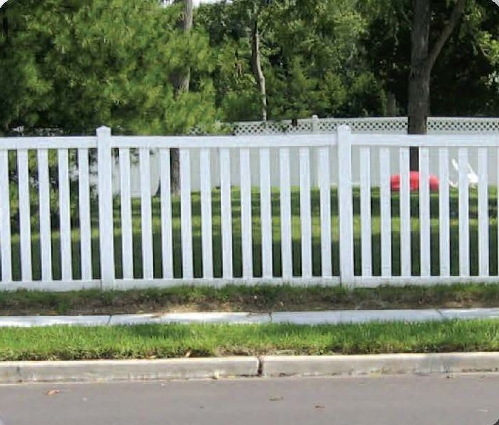 White picket fence bordering a green lawn, next to a road with trees in the background.