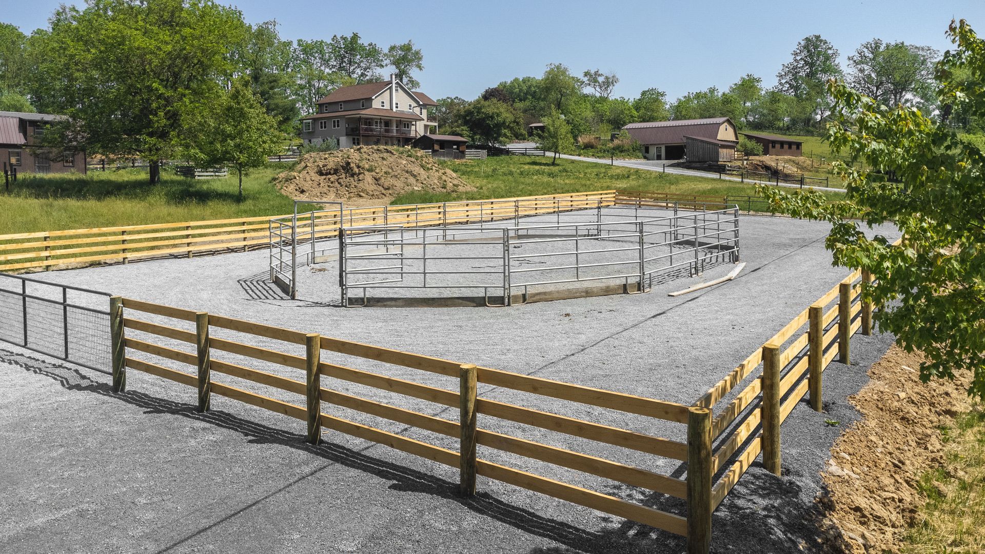 Construction site with circular foundation surrounded by gravel and wooden fences, with a house and trees in the background.