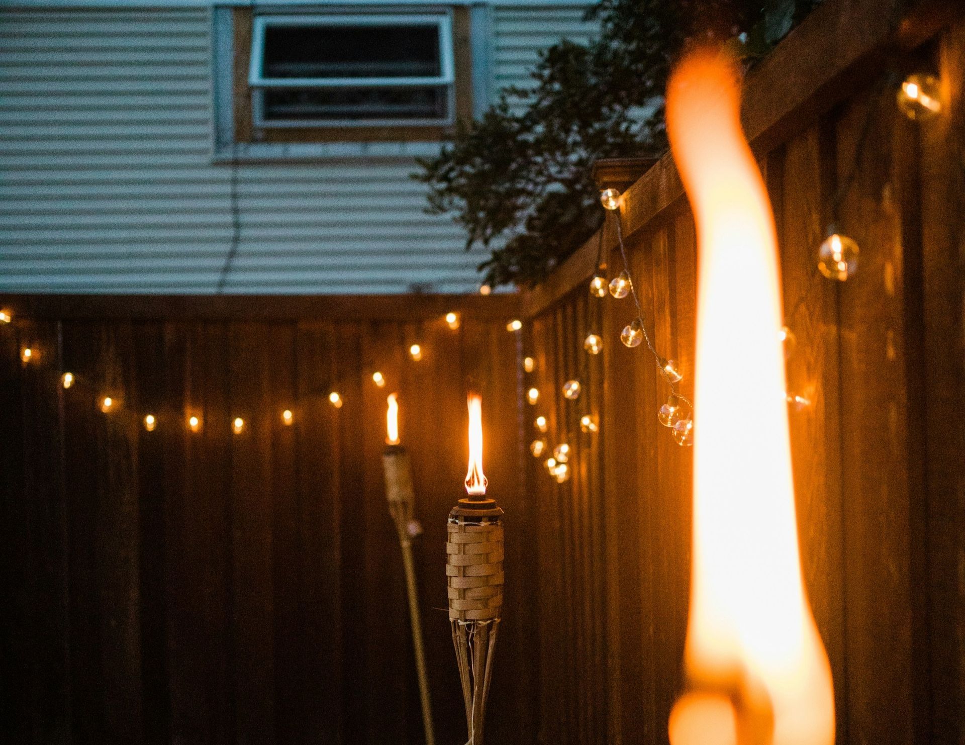 String lights and tiki torches glow along a wooden fence at night, with a house window above.
