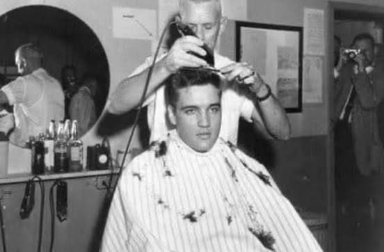 Elvis Presley getting a haircut in a barbershop; barber using clippers.