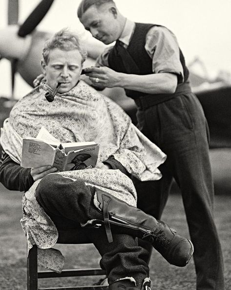 Man getting a haircut while reading and smoking a pipe, near an airplane. A second man is cutting the hair.
