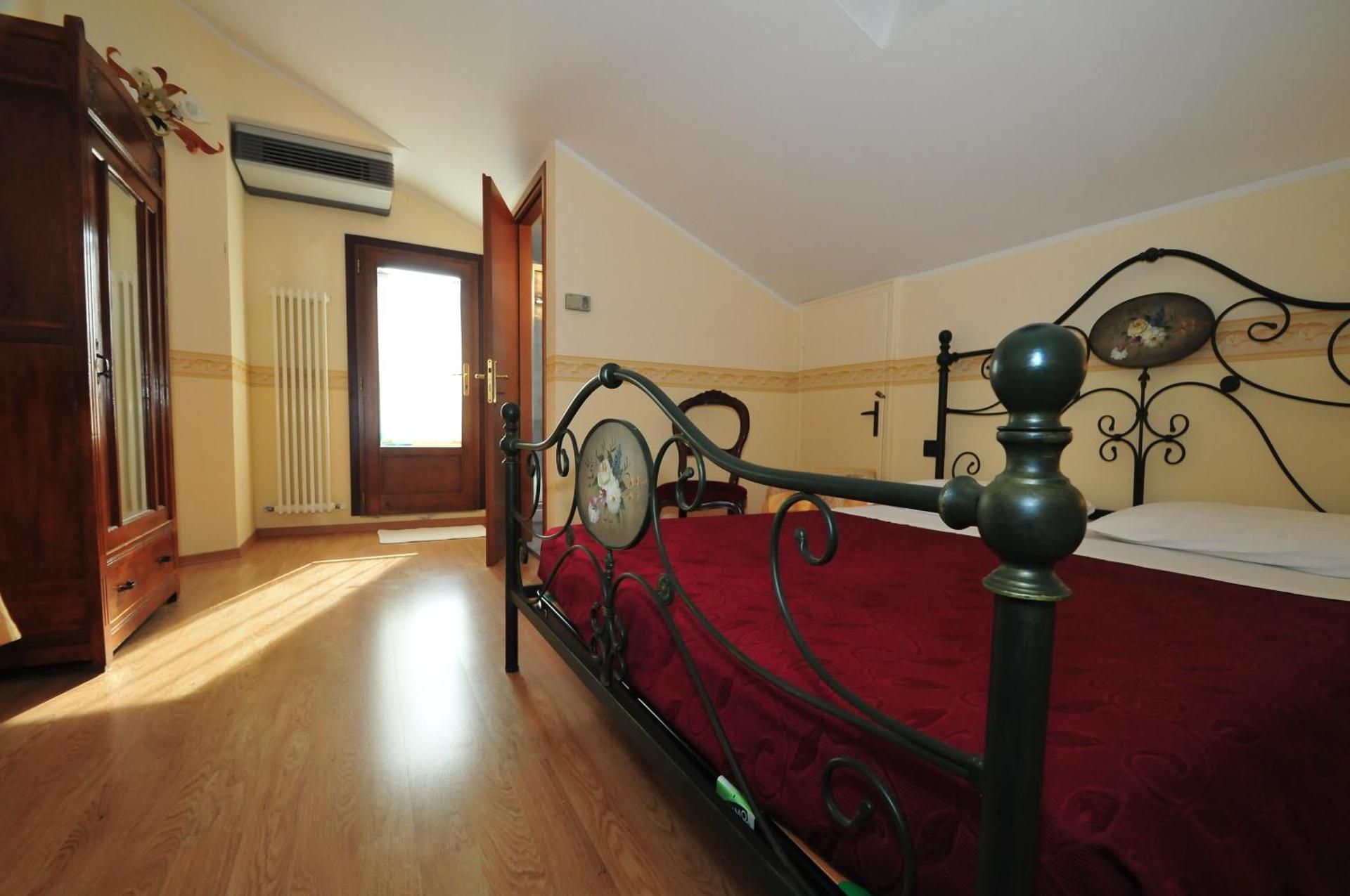 Bedroom with wrought-iron bed, red bedspread, wooden floor, and sunlight from a window and open balcony door