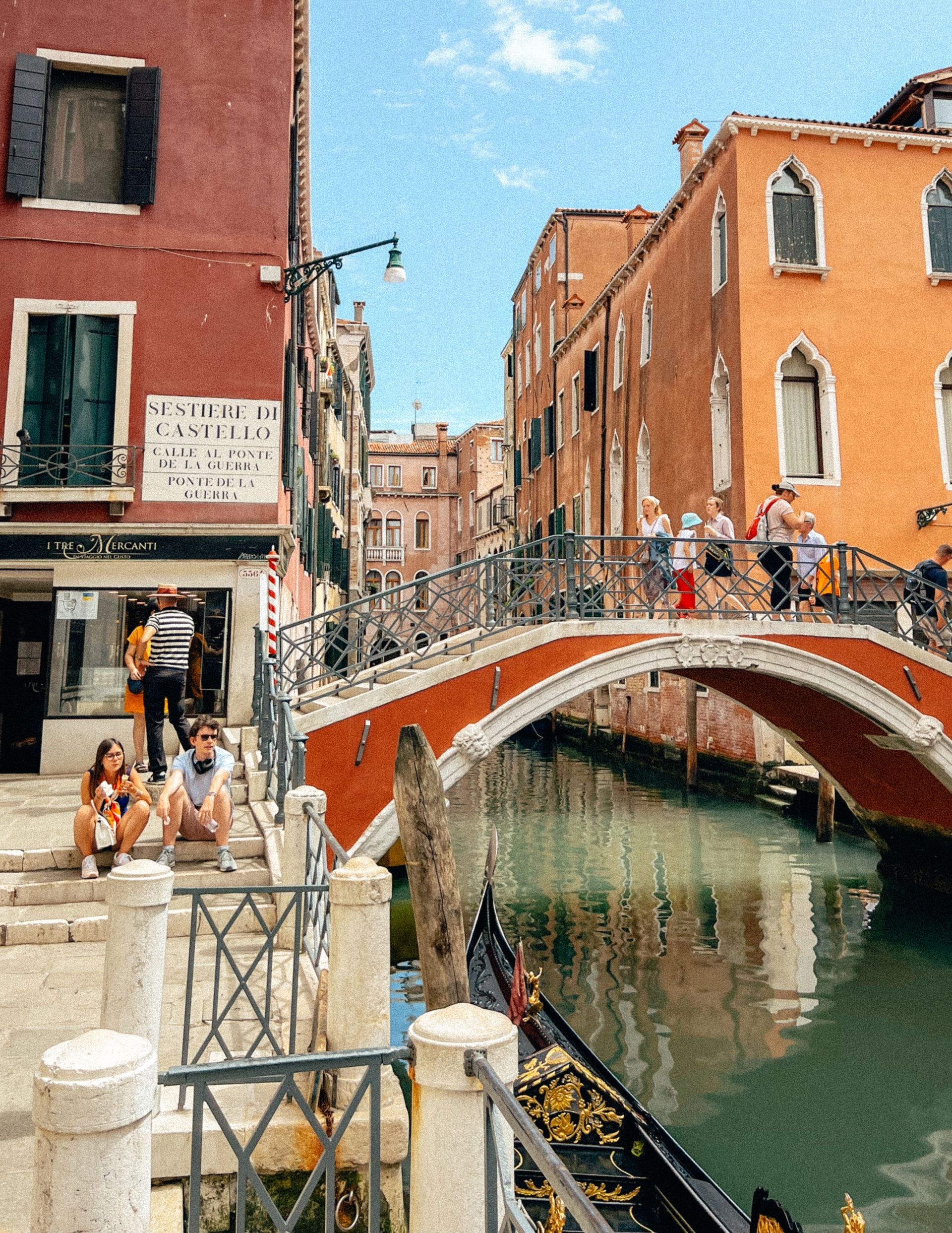 Venice canal with a red arched bridge, colorful buildings, and tourists walking along the waterway