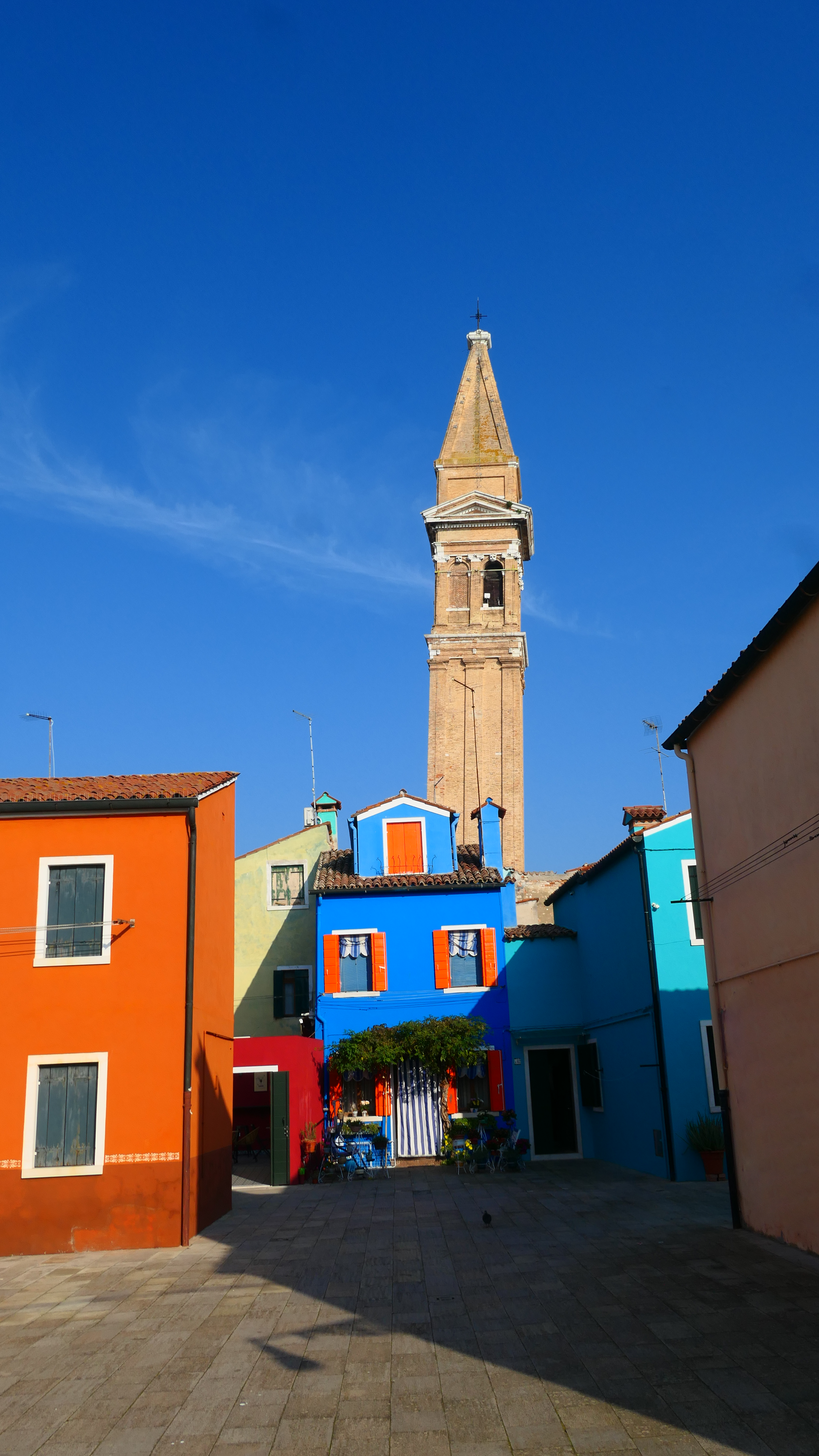 Colorful houses in a sunny square with a tall stone bell tower against a blue sky