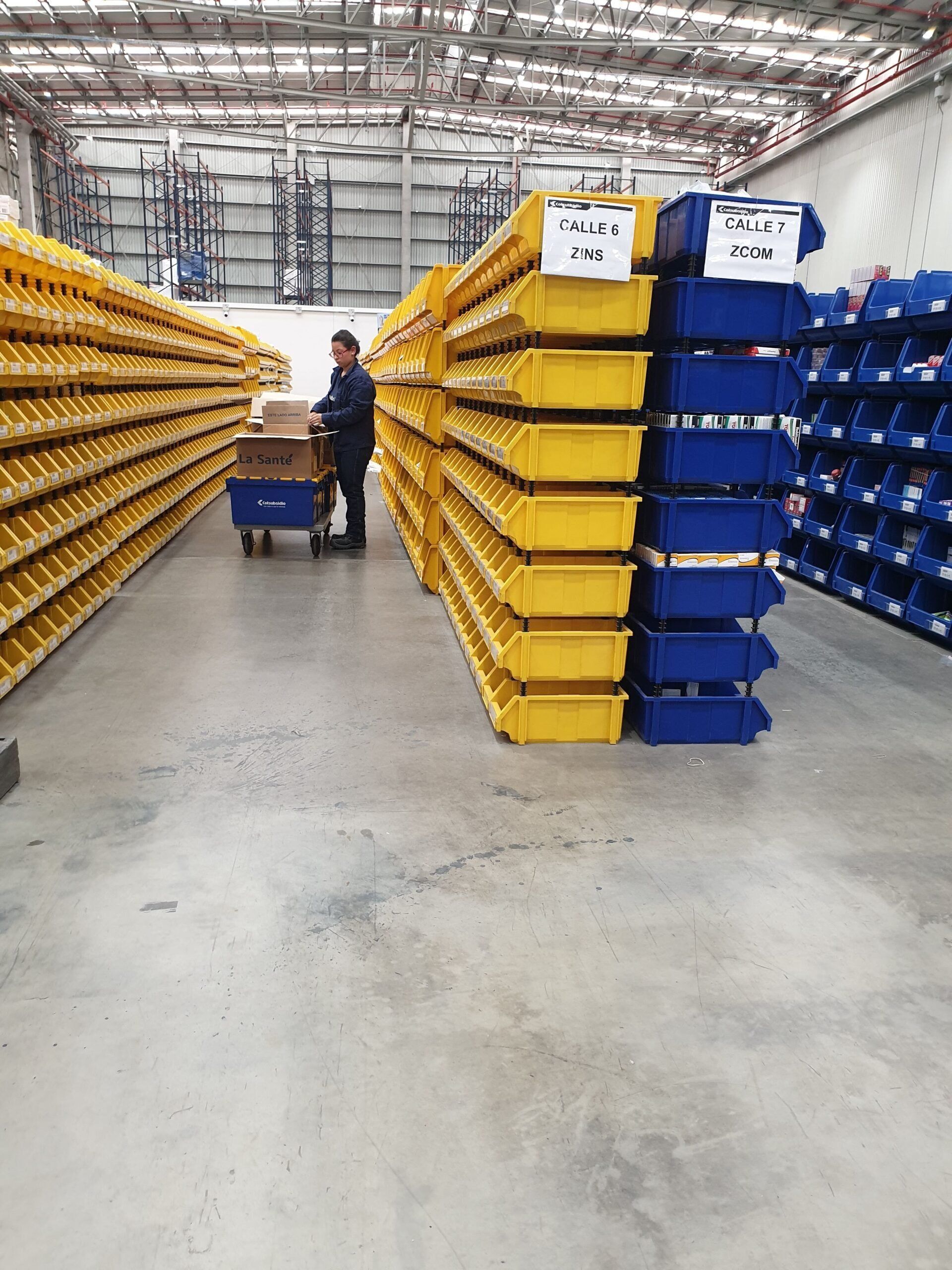 Un hombre empuja un carrito en un almacén lleno de contenedores amarillos y azules.