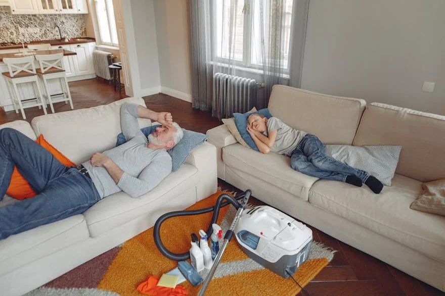 Two people relaxing on sofas in a living room while a vacuum cleaner sits on the rug.
