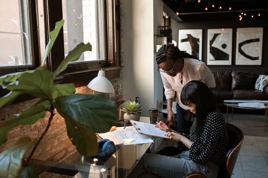 Two people reviewing papers at a café table beside a large window and a leafy plant