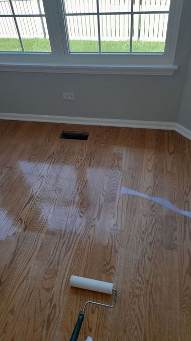 Wooden floor being refinished with a paint roller near a window and vent.
