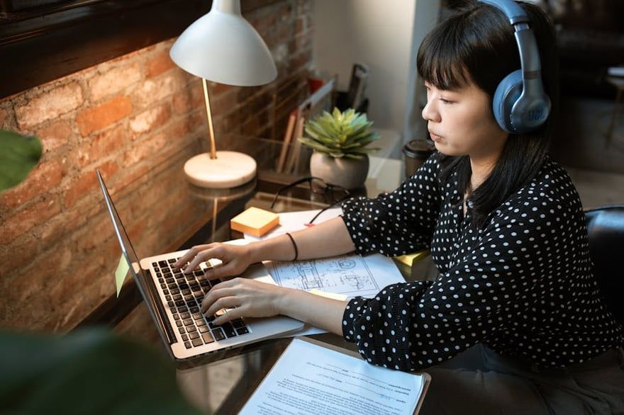Person wearing headphones types on a laptop at a desk beside a lamp and notebook.