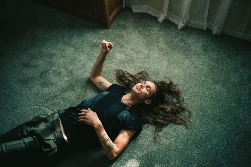 Woman lying on green carpet near a window, one arm raised, with a pensive expression