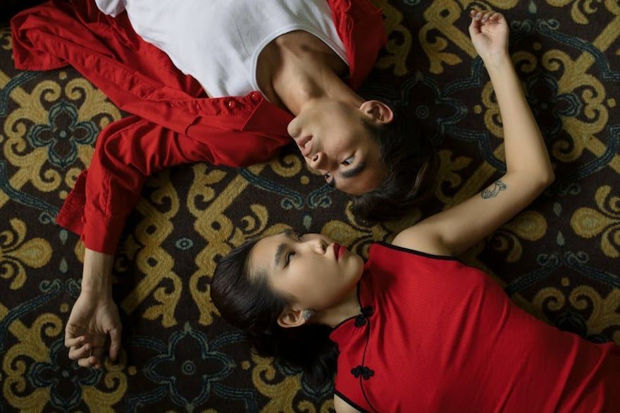 Two people in red tops lying on a patterned rug, facing each other and holding hands.