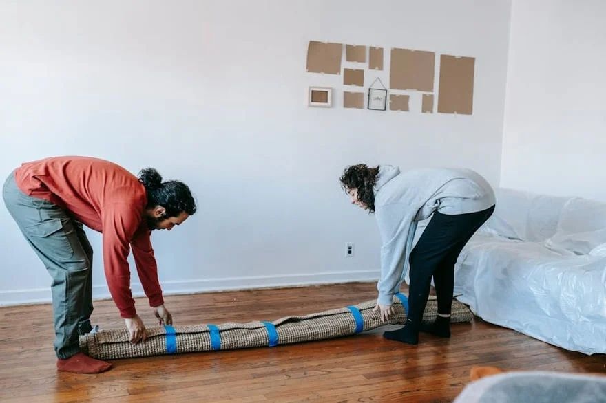 Two people unrolling a carpet in a bare room with white walls and hardwood floors