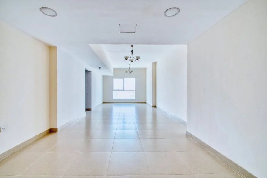 Bright empty living room with white walls, tiled floor, ceiling lights, and a window at the far end