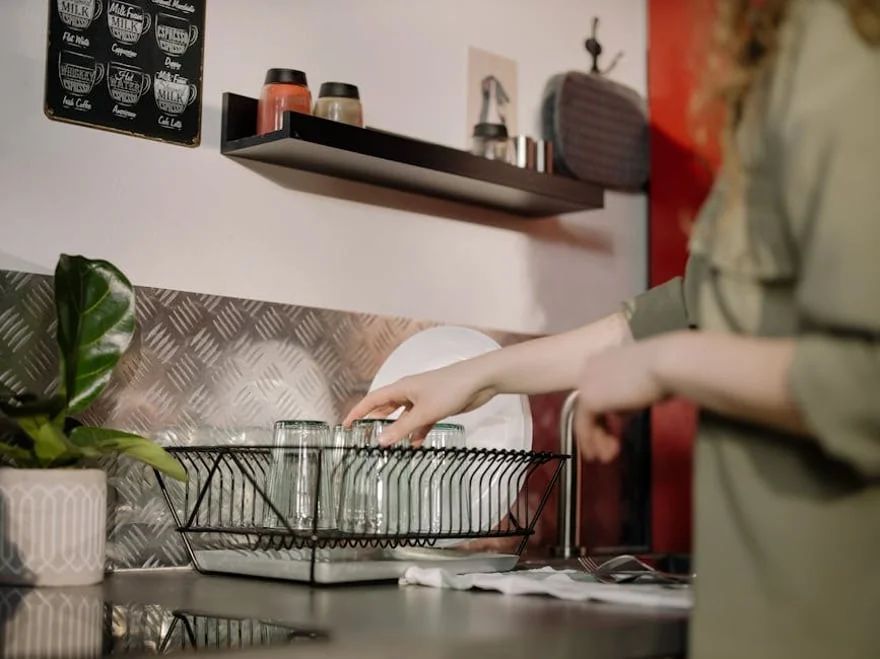 Person loading a wire dish rack in a kitchen with a plant and shelf in the background