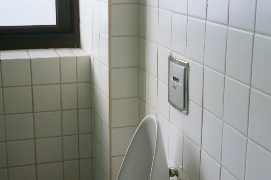 Tiled bathroom corner with a toilet, wall-mounted flush panel, and a small window.