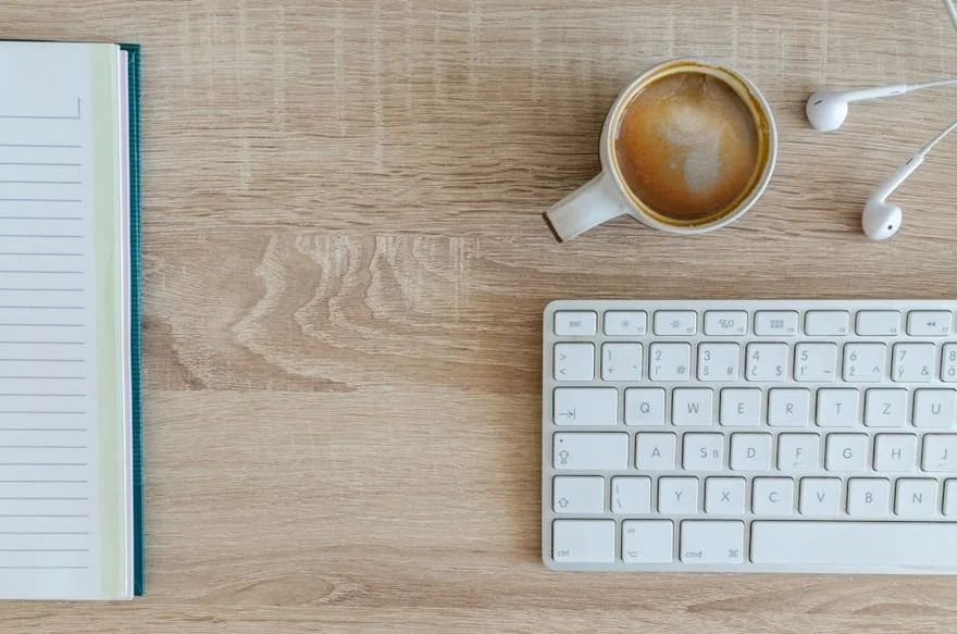 Desk with notebook, coffee cup, earbuds, and white keyboard on a wooden surface.