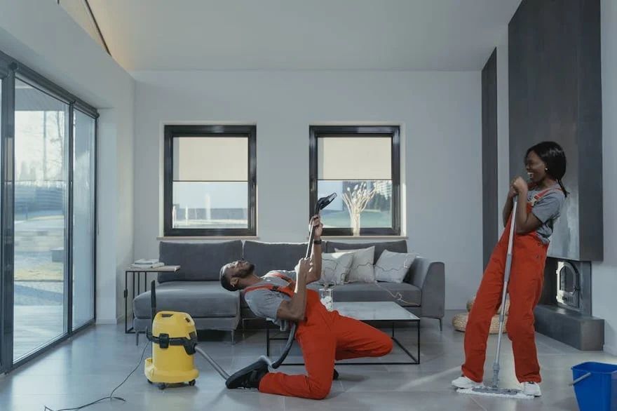 Two workers in orange overalls cleaning a bright modern living room with a vacuum and spray bottle