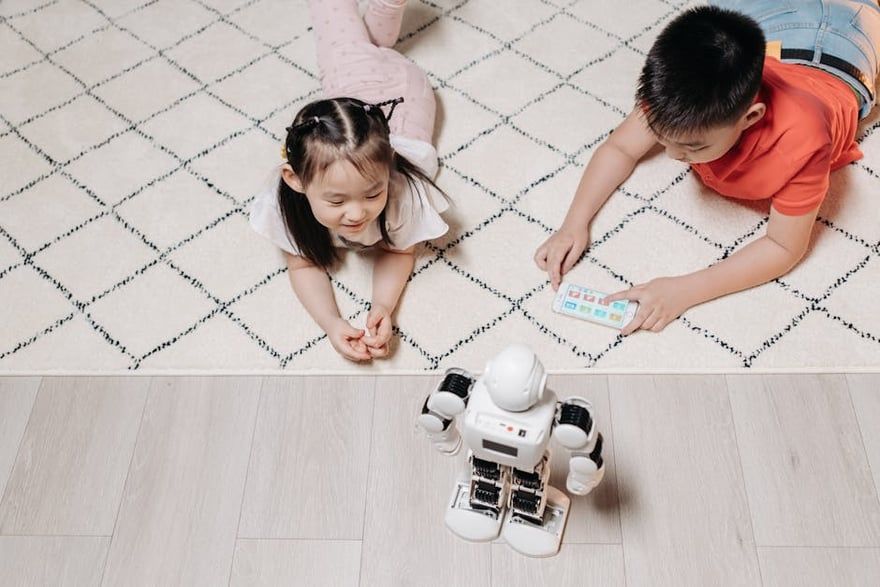 Two children lying on a rug, watching a small white robot toy on the floor
