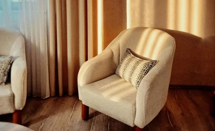 Beige armchair with patterned pillow beside curtains in a softly lit room