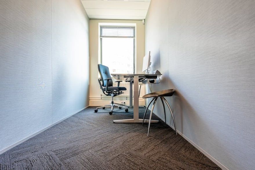 Empty office hallway with a desk, chair, and computer near a window.