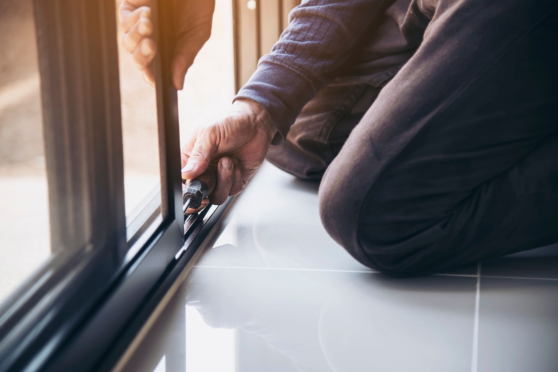 A man is kneeling on the floor fixing a window with a screwdriver.