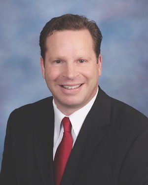 Portrait of a smiling man in a black suit, white shirt, and red tie against a blue background